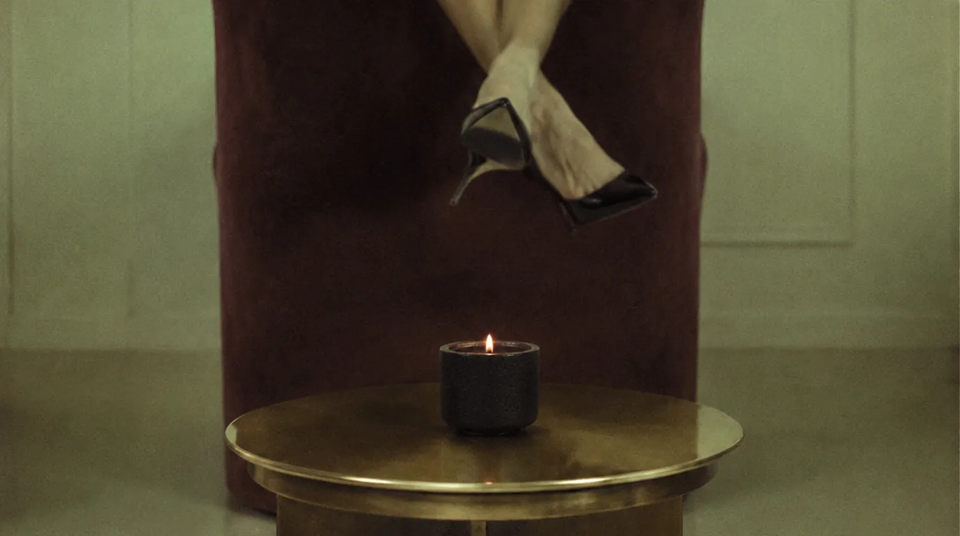 A single black candle burning on a long dark stone dining table in a brutalist dining room, the flame is the only light source